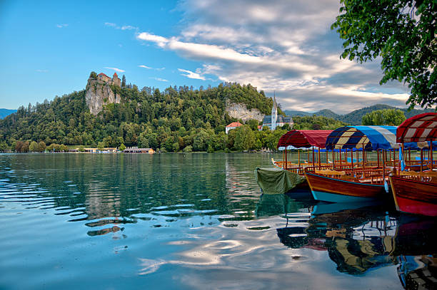 Traditional wooden pletna boats on the shores of Lake Bled in Slovenia, with Bled Castle overlooking the lake from a forested cliff under a partly cloudy sky.